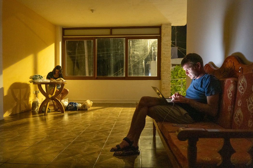 Un uomo è seduto su una panca di legno in un corridoio illuminato dal sole e guarda il suo smartphone; sullo sfondo un’altra persona è seduta vicino a un tavolo accanto alla finestra.