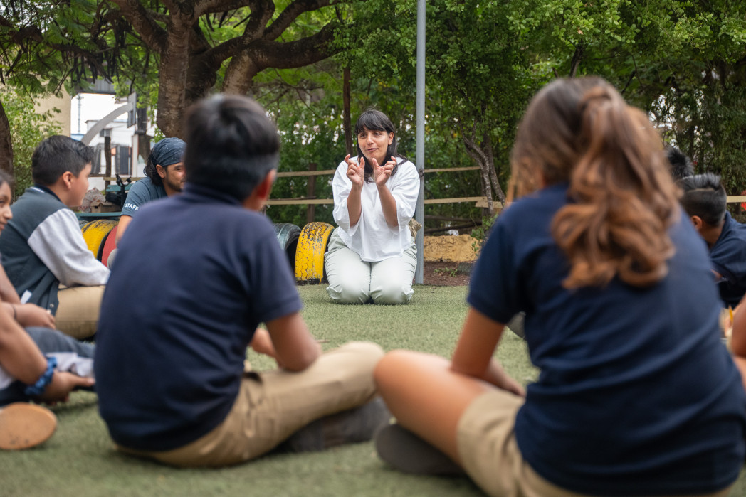 Una donna è seduta all’aperto su un prato e spiega qualcosa usando gesti delle mani, mentre alcuni bambini siedono in cerchio intorno a lei.