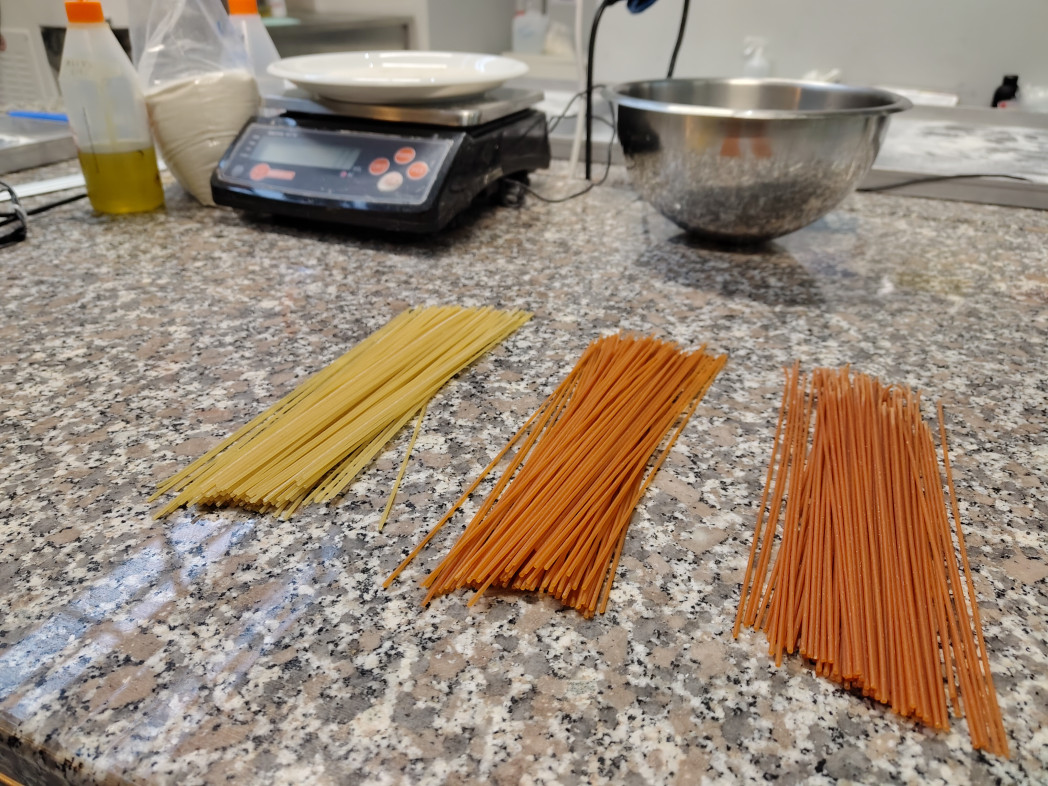 Tre mazzetti di spaghetti crudi di colori diversi sono disposti affiancati su un piano di lavoro; sullo sfondo si vedono una bilancia e delle ciotole.