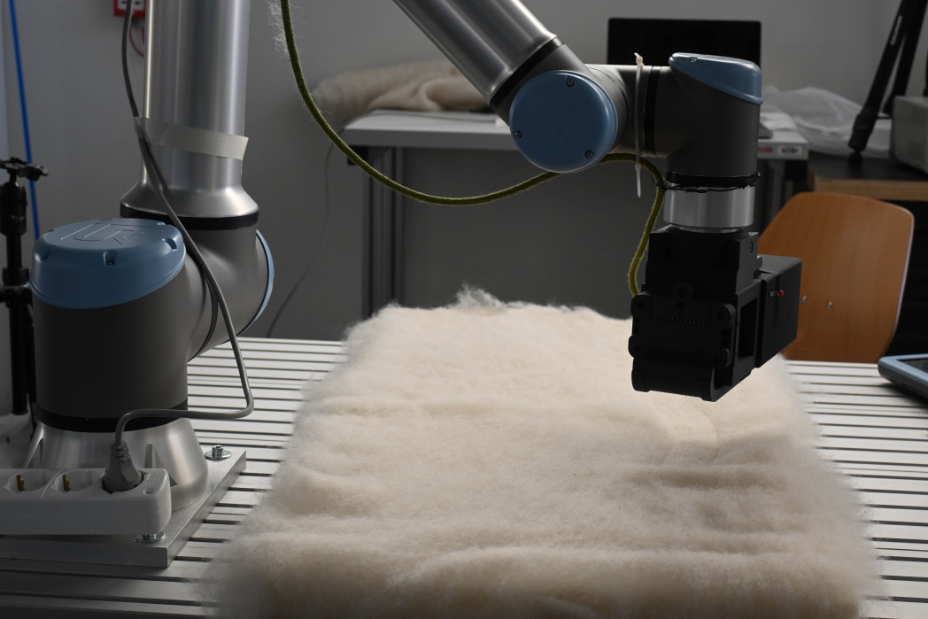 Zwei kollaborative Roboterarme arbeiten über einer hellen Filzmatte auf einem Arbeitstisch in einer Laborumgebung.