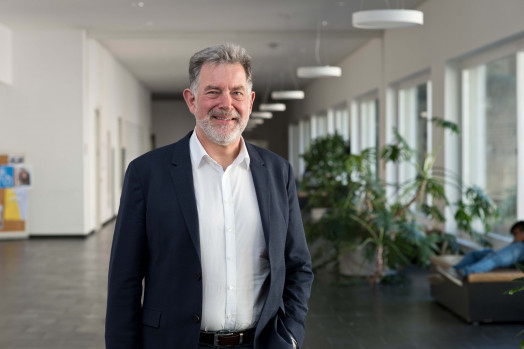 Proträtsfoto von Professor Thomas Bausch in weißem Hemd und schwarzem Sakko, im Hintergrund ein Gang des Campus Bozen der Freien Universität Bozen.