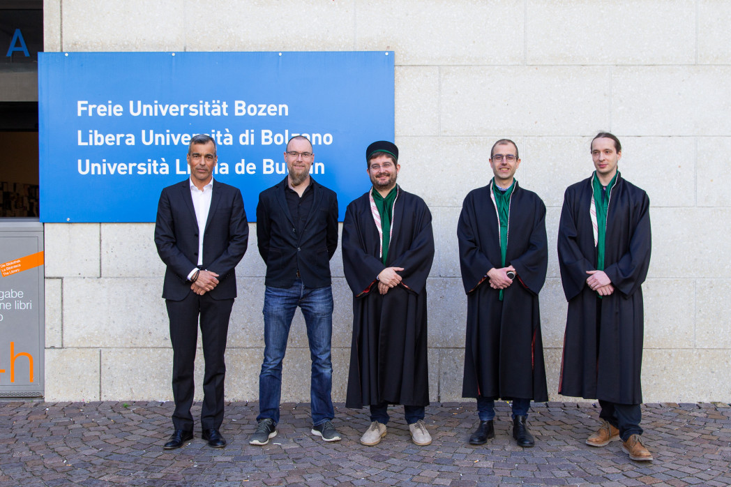 Fünf Personen stehen vor dem Hauptgebäude der Universität in Bozen. Drei davon tragen eine Robe.