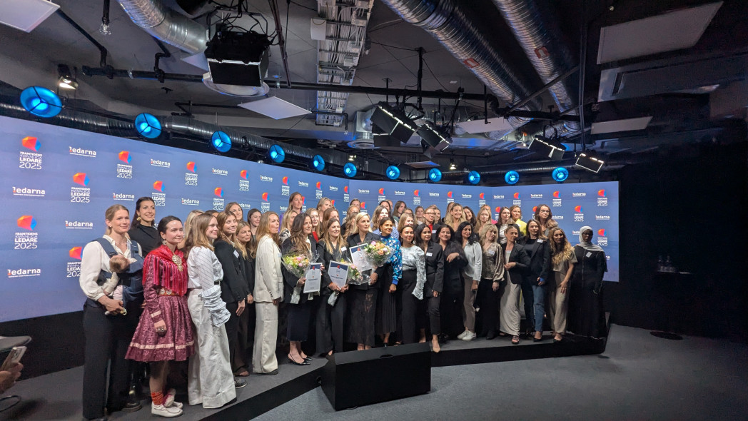 A large group of women pose on a stage in front of a panel with logos, some holding flowers and certificates — the atmosphere is that of an award ceremony or celebration.