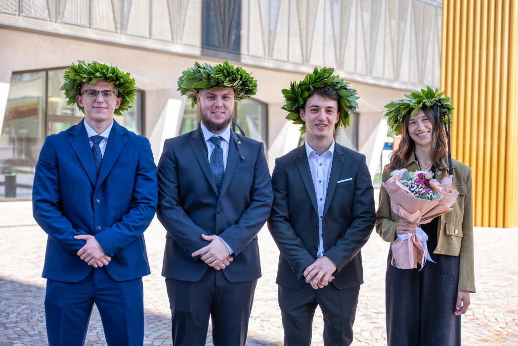 Quattro neolaureati con corona d’alloro e abiti eleganti posano sorridenti davanti all’università; una di loro tiene un mazzo di fiori.