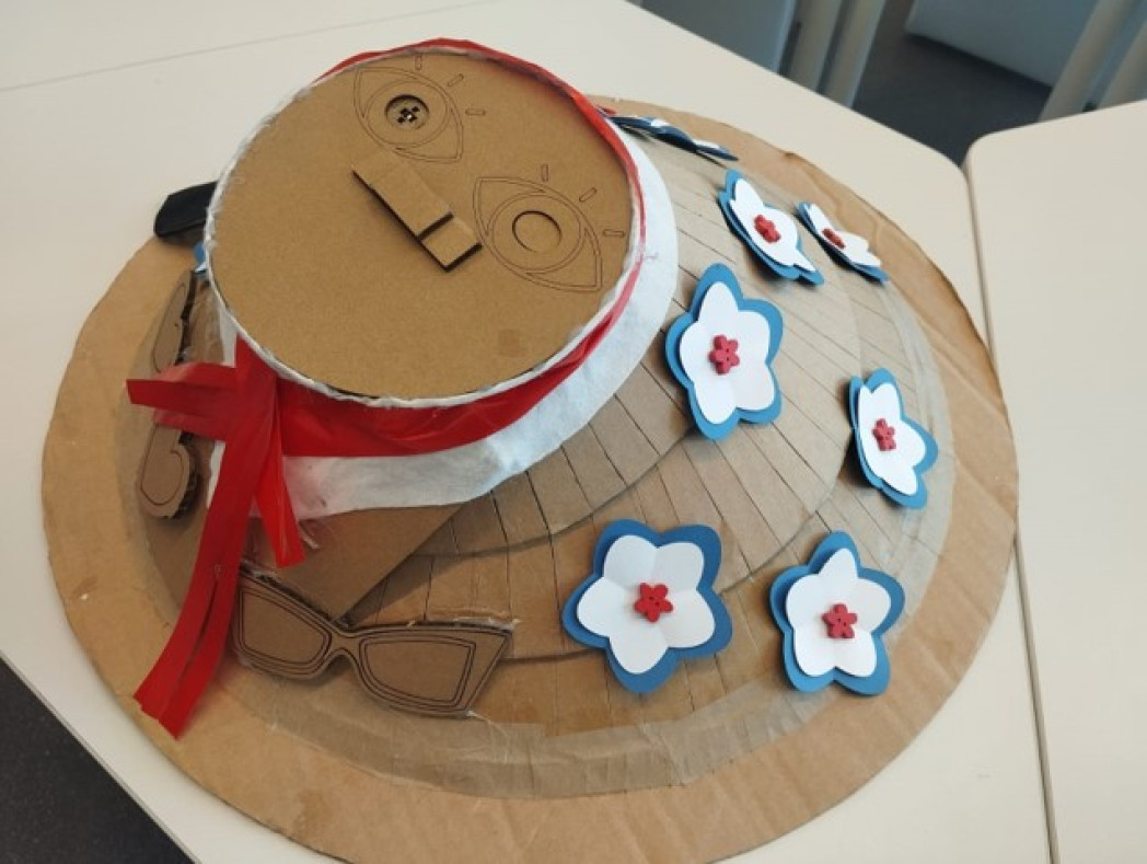 A handmade cardboard hat decorated with flowers, a red ribbon, and sunglasses.