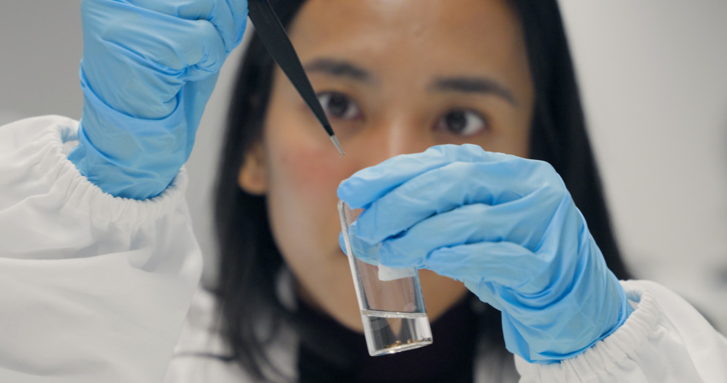 Forscherin im Laborkittel und mit Handschuhen hält ein Reagenzglas mit Flüssigkeit in der Hand.