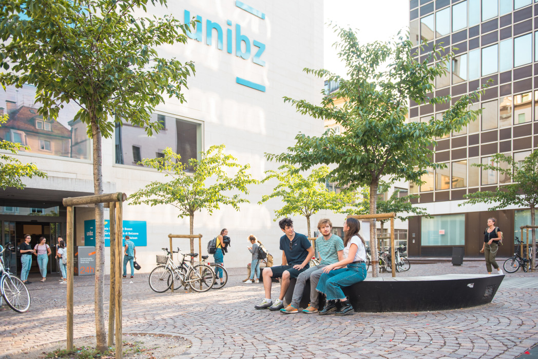 Platz mit Bäumen und Universitätsgebäude im Hintergrund, auf einer Bank und vor Eingang sieht man junge Menschen