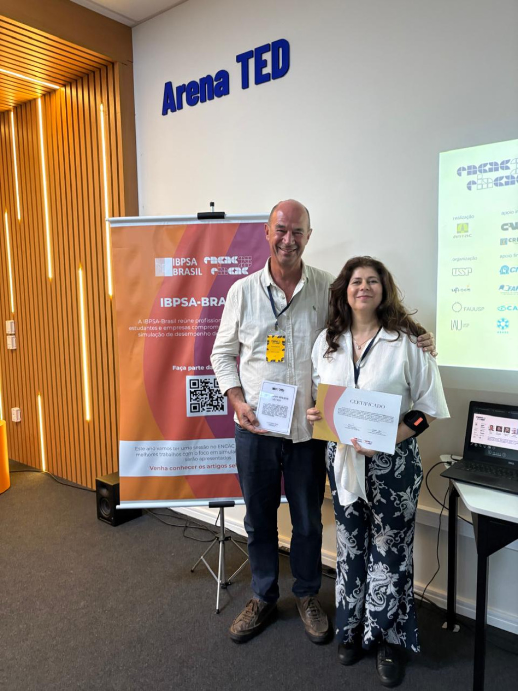 Man and woman standing together holding a certificate in front of Arena TED branded banner and presentation screen.