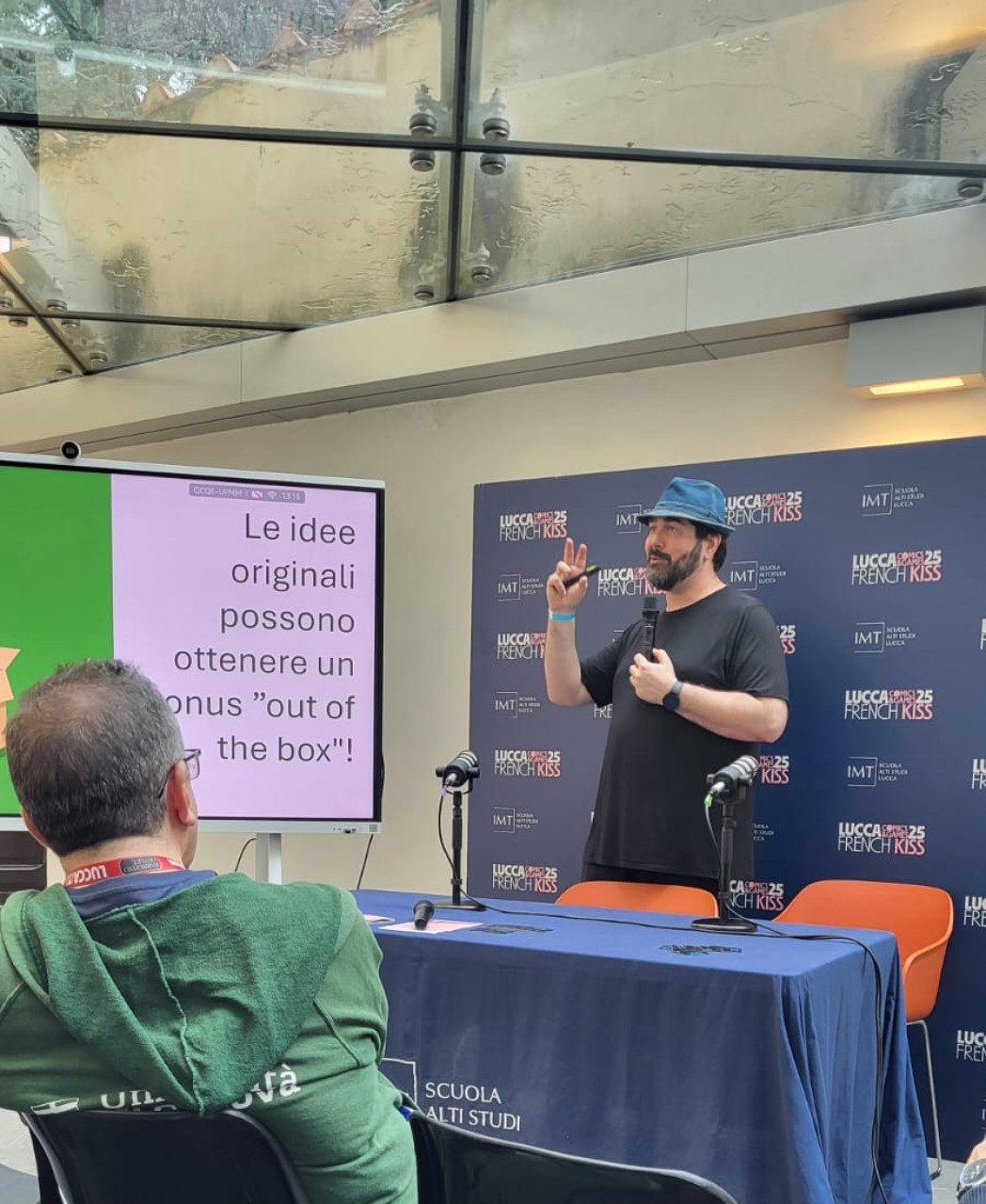 Uomo con cappello tiene un intervento davanti a pubblico e schermo con testo italiano.