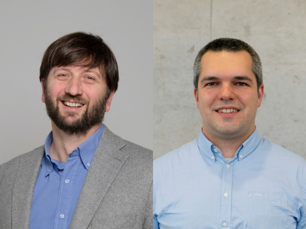 Due uomini affiancati davanti a uno sfondo neutro guardano la fotocamera sorridendo; a sinistra con barba e giacca grigia, a destra senza barba con camicia azzurra.