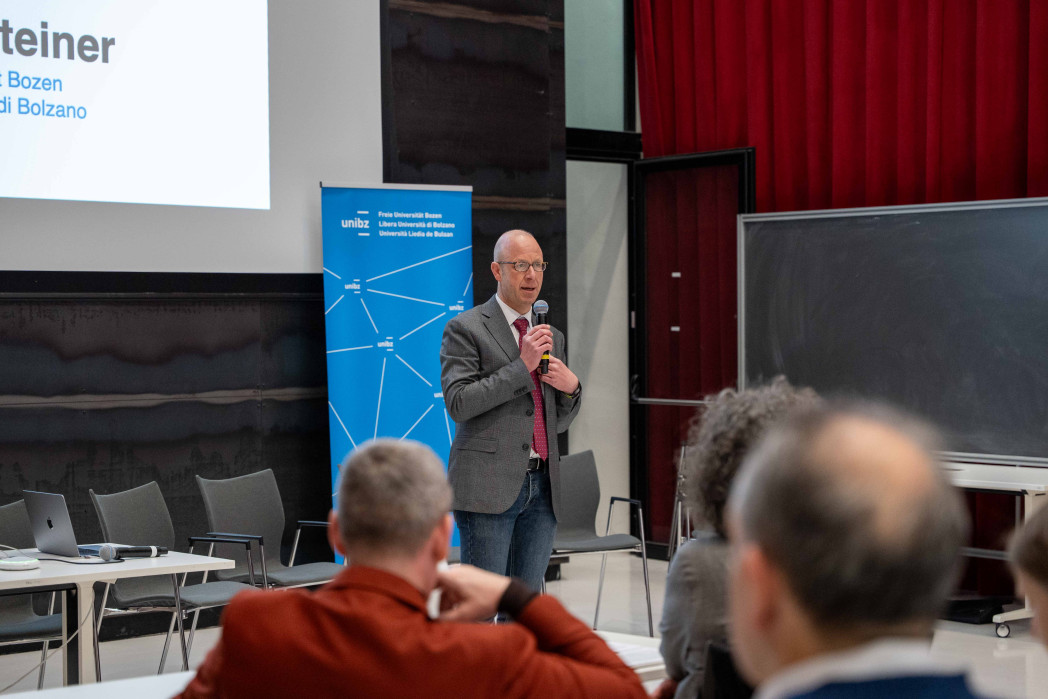 Un uomo con un microfono parla in un’aula davanti al pubblico; sullo sfondo si vedono una diapositiva di presentazione, un banner dell’università e una lavagna.