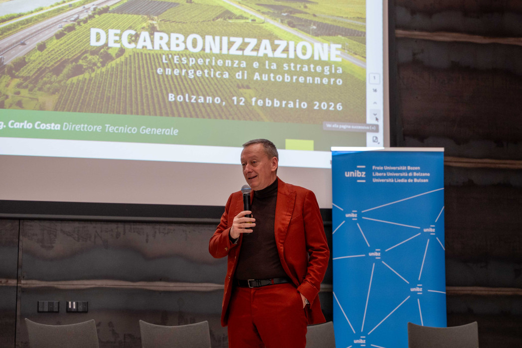 Un uomo in abito rosso parla con un microfono davanti a una diapositiva intitolata “Decarbonizzazione – L’esperienza e la strategia di Autobrennero”; accanto si vede un banner dell’università.