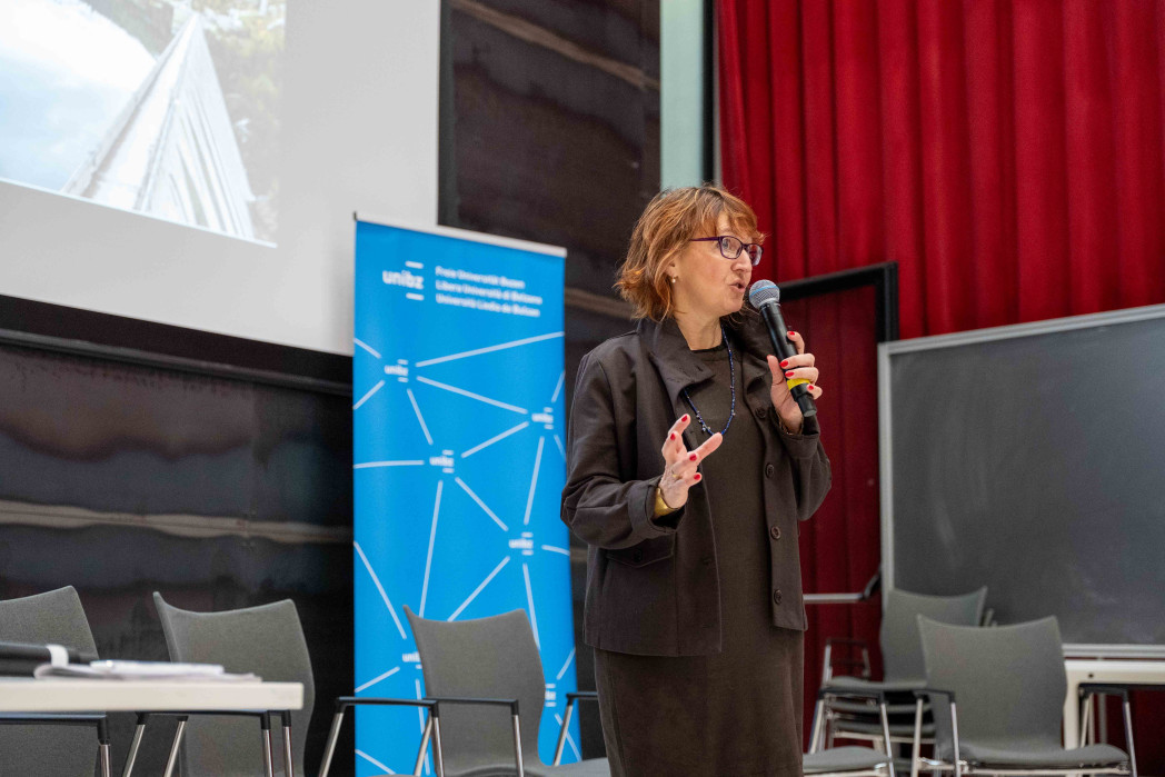 Una donna parla con un microfono in un’aula; sullo sfondo si vedono una diapositiva di presentazione, un banner universitario e una lavagna.