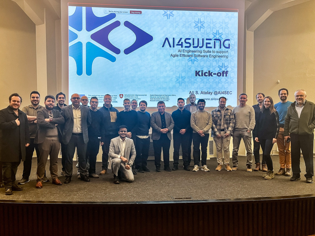 Foto di gruppo di circa venti persone su un palco davanti a un grande schermo con il logo “AI4Synergy” e la scritta “Kick-off”.