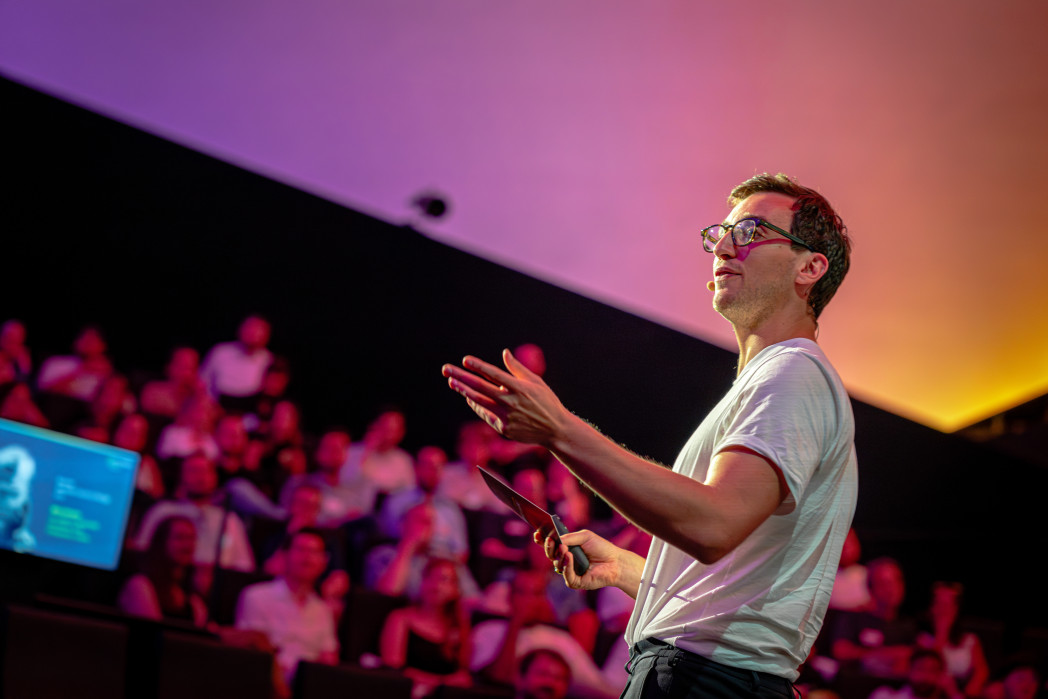 Vortragender in weißem T-Shirt spricht vor Menschenmenge