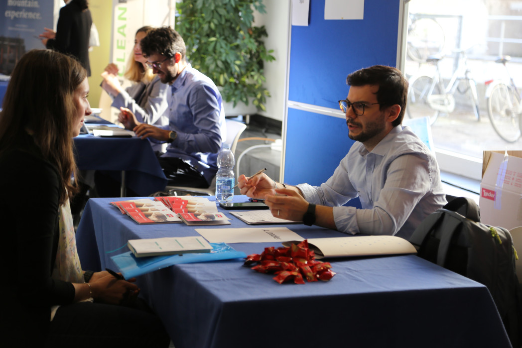 Tisch an dem sich ein Mann und eine Frau gegenübersitzen, im Hintergrund ein weiterer Mann. 