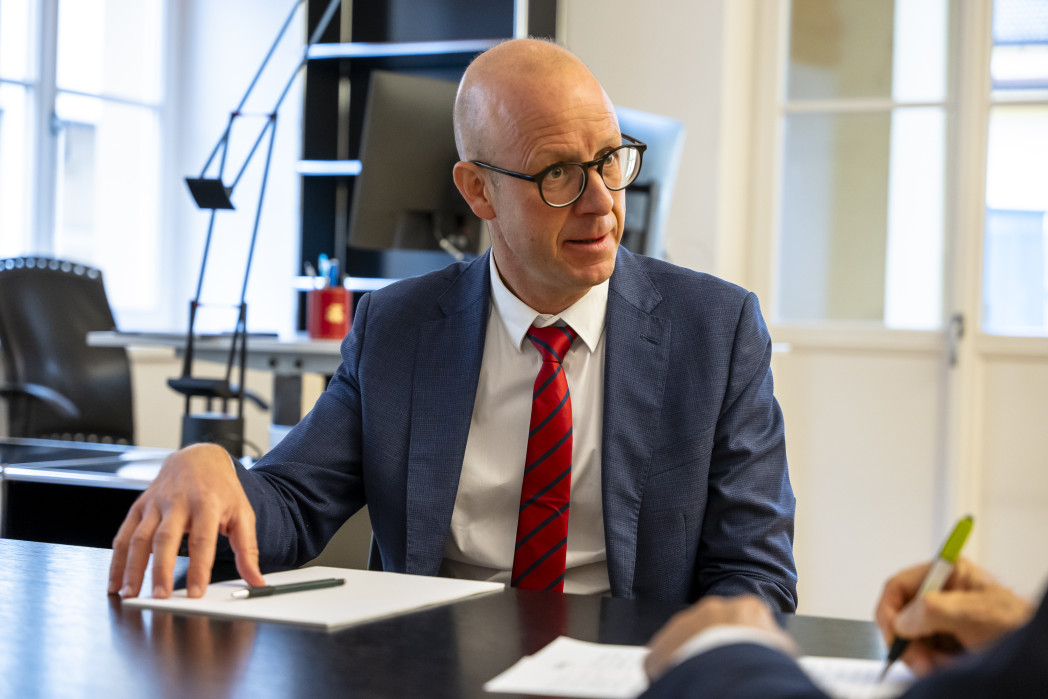 Mann mit Sakko und roter Krawate an Schreibisch, der mit jemanden spricht und mit linker Hand gestikuliert