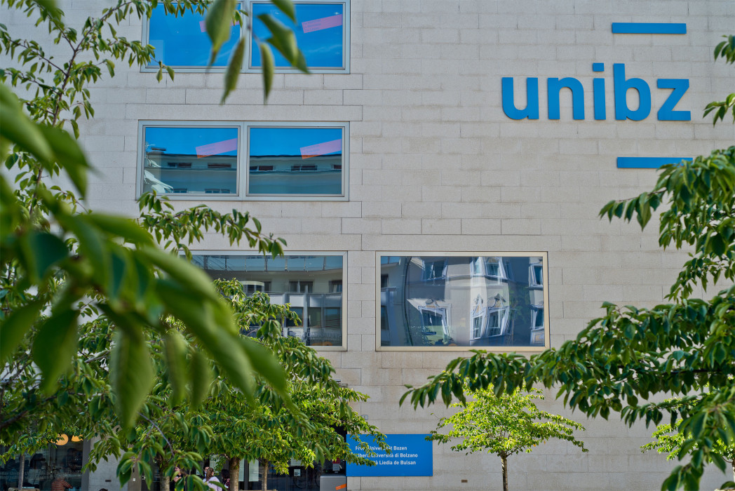 Fassade der unibz mit blauem Logo auf heller Wand, umgeben von grünen Blättern im Vordergrund