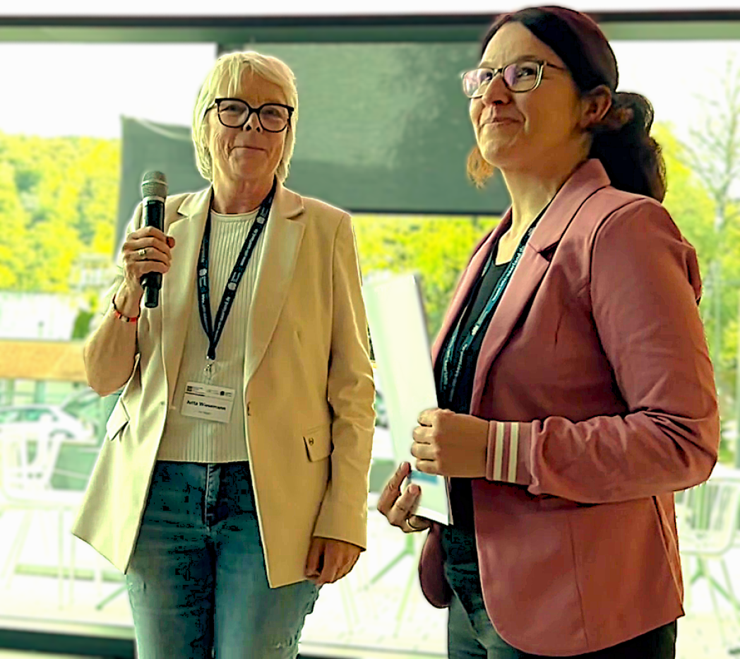 Eine Frau mit Mikrofon und eine Frau mit Urkunde in der Hand auf einem Podium