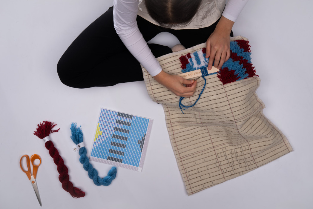 Una persona seduta per terra e fa i nodi. Accanto a lei ci sono filati, forbici e un modello.