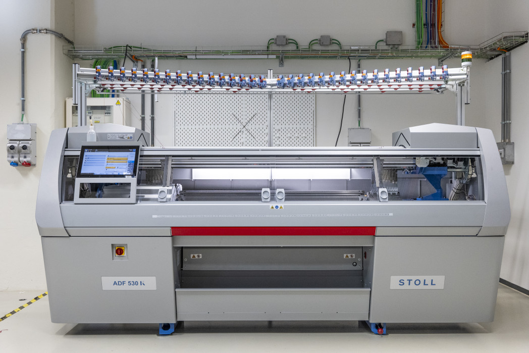 Eine große Industriestrickmaschine steht in einem Raum. An der Wand dahinter sind einige Schalter und Kabelkanäle angebracht