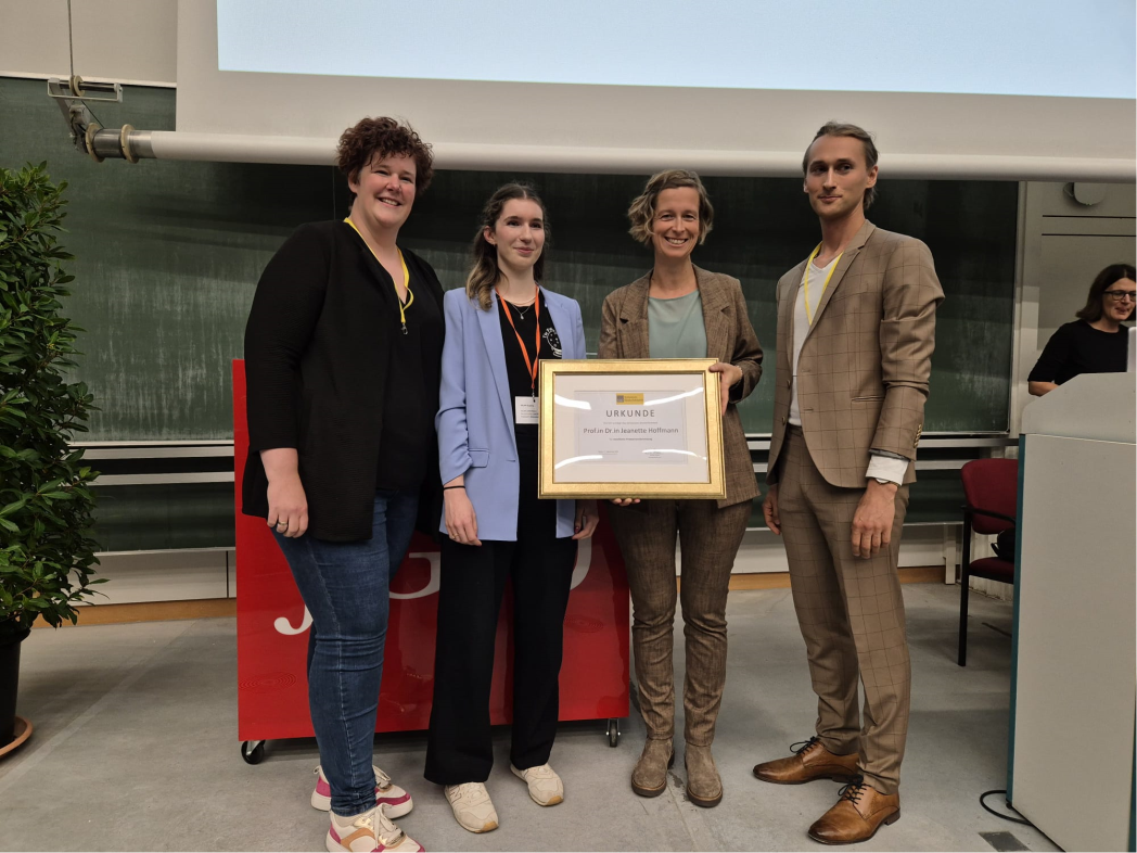 Gruppenbild von vier Personen, eine hält gerahmte Auszeichnung in die Kamera