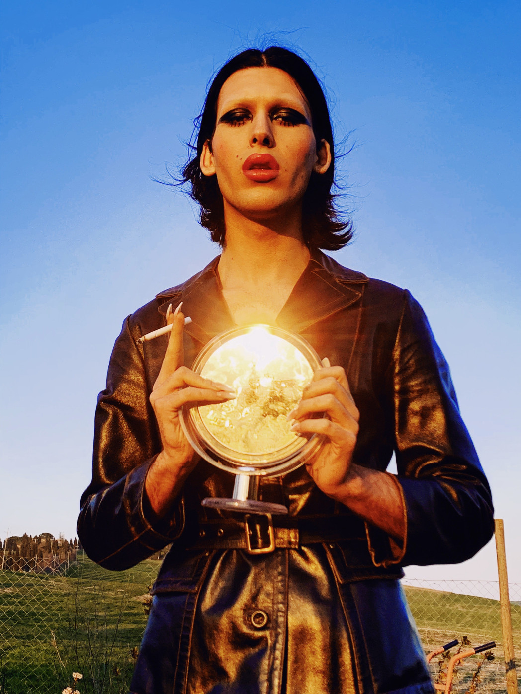 Eine stark geschminkte Person mit langen Fingernägeln hält einen Schminkspiegel in beiden Händen. In einer Hand hält sie noch eine Zigarette.