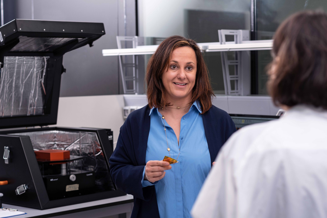 Una donna si trova in un laboratorio e tiene in mano un piccolo componente, parlando con un’altra persona tra apparecchiature tecniche.