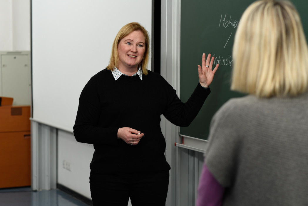 Professorin Gunkel steht vor einer Tafel und erklärt etwas einer gegenüberstehender Frau, die man nur von hinten sieht. 