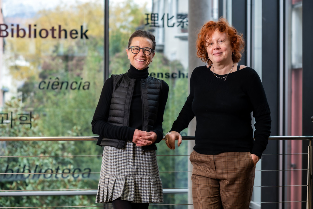 Die Professorinnen Andrea Abel und Birgit Alber sind an ein Geländer gelehnt. Im Hintergrund eine Glasscheibe durch die Bäume sichtbar sind.