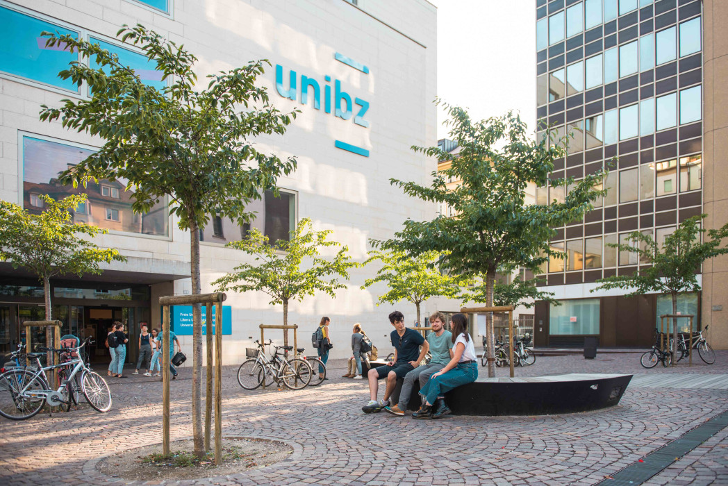 Una piazza con alberi. Sullo sfondo l'edificio della Libera Università. L'edificio è contrassegnato dalla scritta unibz.